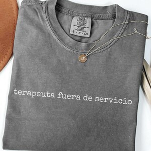 May include: A charcoal gray short-sleeve t-shirt with a crew neckline and the white text "terapeuta fuera de servicio" printed across the chest. The shirt features a Comfort Colors brand label inside the collar and a thin gold-tone necklace.