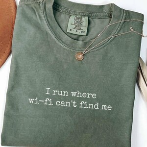 May include: A moss green short-sleeve t-shirt with a crew neckline and the white embroidered text I run where wi-fi can't find me. The shirt features a Comfort Colors brand label at the inner neck and is paired with a thin gold-tone chain necklace.