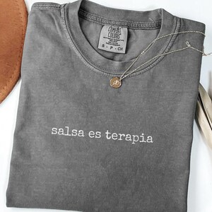 Puede incluir: Una camiseta gris carbón de manga corta con cuello redondo y el texto bordado en blanco "salsa es terapia" centrado en el pecho. La prenda incluye una etiqueta de la marca Comfort Colors en el cuello interior y un collar de cadena dorada.