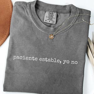 May include: A charcoal gray short-sleeve t-shirt with a crew neckline. The shirt features the white text "paciente estable, yo no" printed across the chest. A thin gold-toned chain necklace with a small circular pendant rests on the shirt.
