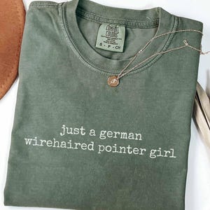 May include: A moss green short-sleeve t-shirt with a crew neckline and the white text "just a german wirehaired pointer girl" printed on the front. The shirt features a Comfort Colors brand label at the inner neck and a thin gold-tone chain necklace.