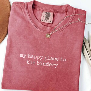 May include: A crimson short-sleeved t-shirt with a crew neckline and the white embroidered text 'my happy place is the bindery' centered on the chest. The shirt features a Comfort Colors brand label at the inner neck and a size small tag.