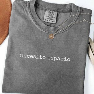 May include: A charcoal gray short-sleeve t-shirt with the white embroidered text "necesito espacio" across the chest. The shirt features a crew neckline and a Comfort Colors brand label inside the collar, paired with a delicate gold-tone necklace.