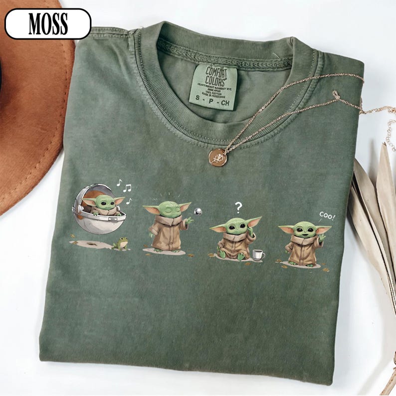 May include: Green Comfort Colors t-shirt featuring a graphic of Grogu in various poses. The design includes Grogu in a pod, playing with a ball, with a question mark, and saying "Coo!". A gold necklace and the word "MOSS" are also visible.