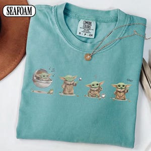 May include: Seafoam green t-shirt featuring a series of illustrations of a character from a popular science fiction franchise. The character is depicted in various poses, with the word "coo!" printed on the shirt.