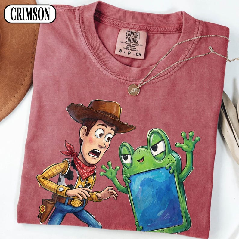 May include: A crimson short-sleeve t-shirt with a crew neckline and a Comfort Colors brand label. The graphic features a cartoon cowboy character in a brown hat and a green frog-shaped device with a blue screen. The text CRIMSON is visible.