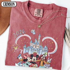 May include: Crimson red t-shirt with a Disney castle graphic, Mickey Mouse, Minnie Mouse, Donald Duck, Goofy, and Daisy Duck. The shirt has the year 2026 printed on it, and a Comfort Colors tag. A gold necklace is draped across the shirt.