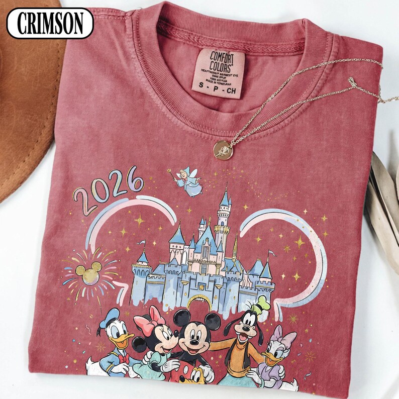 May include: Crimson red t-shirt with a Disney castle graphic, Mickey Mouse, Minnie Mouse, Donald Duck, Goofy, and Daisy Duck. The shirt has the year 2026 printed on it, and a Comfort Colors tag. A gold necklace is draped across the shirt.