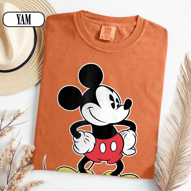 May include: A burnt orange crew neck t-shirt featuring a graphic of Mickey Mouse with black ears, a white face, and red shorts. The shirt has a tag inside the collar that reads Comfort Colors and the size XL. The fabric has a textured appearance.