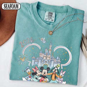 May include: Seafoam green t-shirt with a Disney-themed graphic, including Mickey Mouse, Minnie Mouse, Donald Duck, Daisy Duck, and Goofy, plus a castle and the year 2026. A Comfort Colors label is visible.