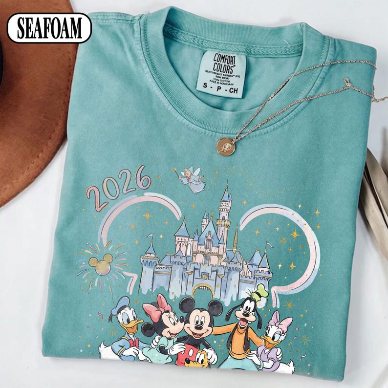 May include: Seafoam green t-shirt with a Disney-themed graphic, including Mickey Mouse, Minnie Mouse, Donald Duck, Daisy Duck, and Goofy, plus a castle and the year 2026. A Comfort Colors label is visible.