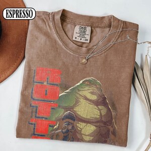 Puede incluir: Camiseta color espresso con un gr&aacute;fico de un alien&iacute;gena verde y la palabra "Hoth" en rojo. La camiseta es de la marca Comfort Colors, talla S-CH. Un collar plateado con un colgante est&aacute; sobre la camiseta.
