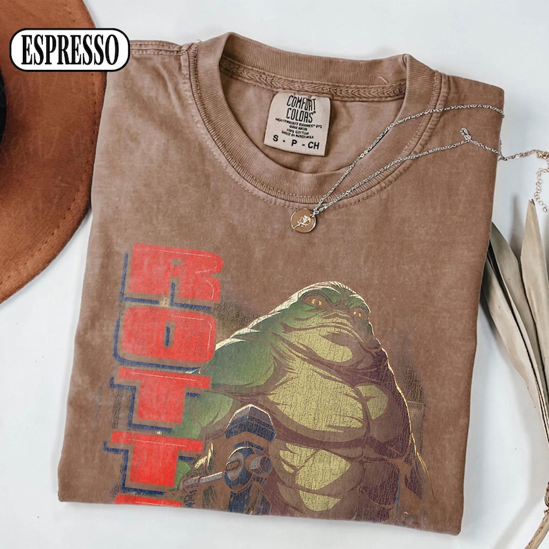 Puede incluir: Camiseta color espresso con un gr&aacute;fico de un alien&iacute;gena verde y la palabra "Hoth" en rojo. La camiseta es de la marca Comfort Colors, talla S-CH. Un collar plateado con un colgante est&aacute; sobre la camiseta.