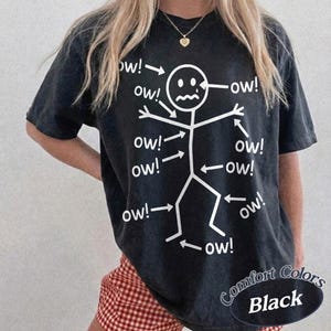 May include: A black short-sleeve t-shirt featuring a white graphic of a stick figure with a pained facial expression. Multiple arrows point to various parts of the stick figure's body, each accompanied by the text "ow!" written in a white font.