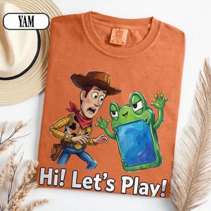 May include: An orange t-shirt featuring a graphic of a cowboy character and a green frog-like creature holding a blue screen. The text "Hi! Let's Play!" is printed in white at the bottom. The shirt has a crew neck and a Comfort Colors brand label.