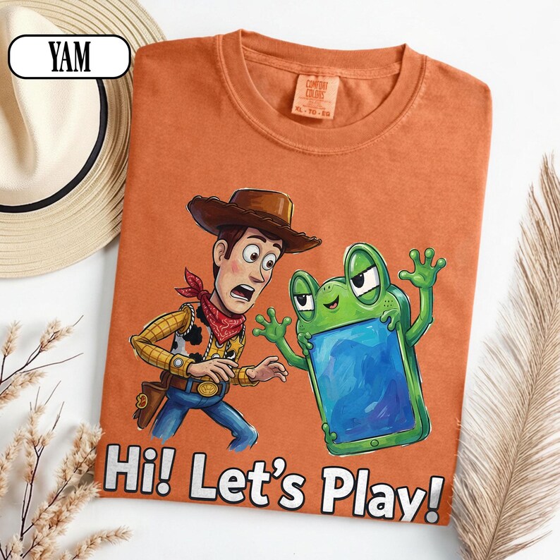 May include: An orange t-shirt featuring a graphic of a cowboy character and a green frog-like creature holding a blue screen. The text "Hi! Let's Play!" is printed in white at the bottom. The shirt has a crew neck and a Comfort Colors brand label.