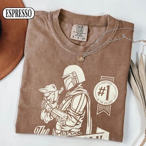 Puede incluir: Camiseta color espresso con un gr&aacute;fico de un personaje sosteniendo una peque&ntilde;a criatura, con el texto "Certified #1 in the Galaxy". La camiseta tiene cuello redondo y mangas cortas. Tambi&eacute;n se ve un collar plateado con un colgante.