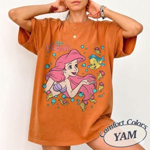 May include: An orange short-sleeve t-shirt featuring a graphic print of Ariel from The Little Mermaid with pink hair, a purple seashell top, and Flounder the fish. The design includes blue bubbles and the text The Little Mermaid in blue.