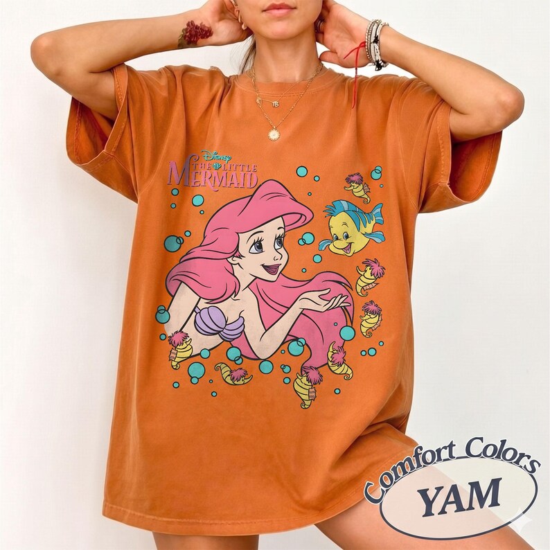 May include: An orange short-sleeve t-shirt featuring a graphic print of Ariel from The Little Mermaid with pink hair, a purple seashell top, and Flounder the fish. The design includes blue bubbles and the text The Little Mermaid in blue.