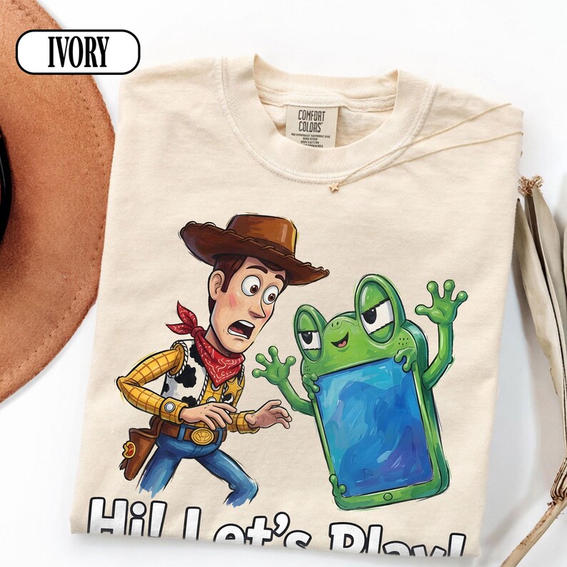 May include: An ivory t-shirt featuring a graphic of a cowboy character with a brown hat and red bandana standing next to a green frog-shaped device with a blue screen. The text "Hi! Let's Play!" is printed at the bottom in black letters.