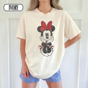 May include: Ivory-colored t-shirt featuring a distressed Minnie Mouse graphic with a red bow. The graphic is centered on the front of the shirt. The shirt has short sleeves and a crew neck.