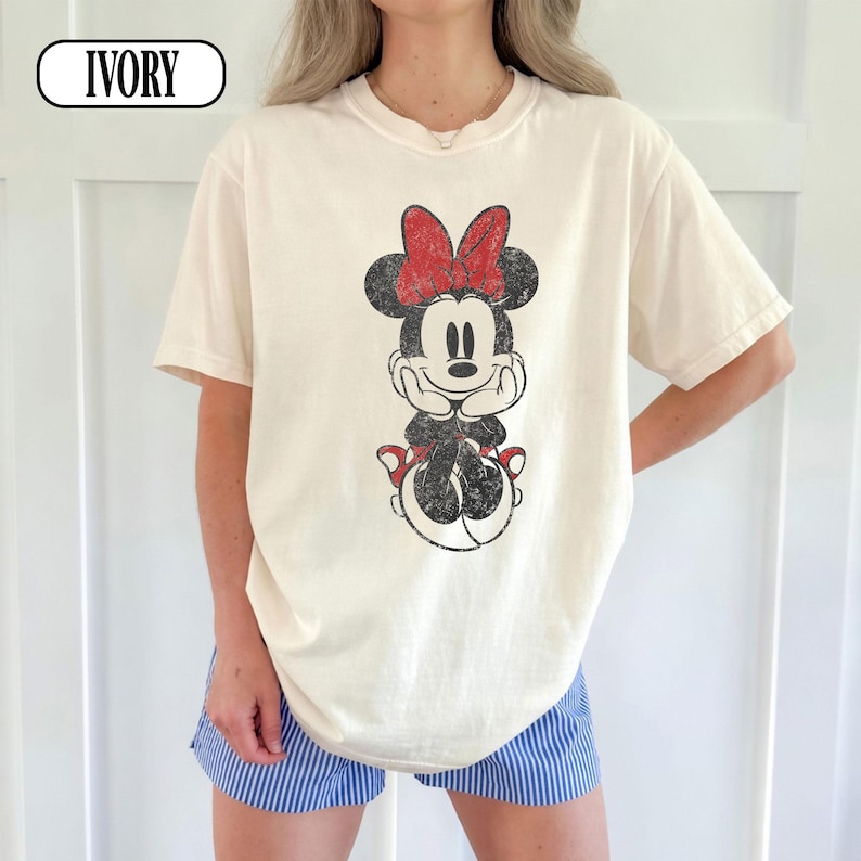 May include: Ivory-colored t-shirt featuring a distressed Minnie Mouse graphic with a red bow. The graphic is centered on the front of the shirt. The shirt has short sleeves and a crew neck.