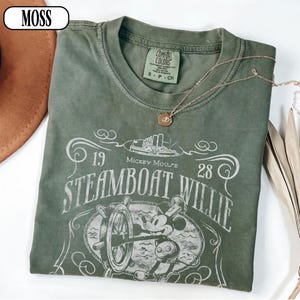 May include: A moss green short-sleeve t-shirt featuring a white graphic print of Mickey Mouse at a ship's wheel. The text reads 1928, Mickey Mouse, and Steamboat Willie. The shirt has a crew neckline and a Comfort Colors brand label inside.