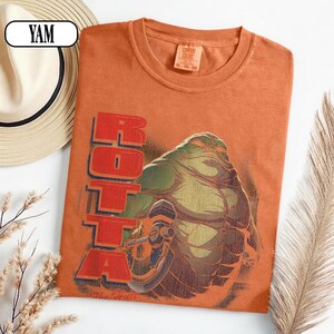 Puede incluir: Camiseta naranja con un gr&aacute;fico de un alien&iacute;gena verde y la palabra "ROTTA" en rojo. La camiseta tiene cuello redondo y una etiqueta "Comfort Colors". Un sombrero de paja y una planta seca est&aacute;n en el fondo. Talla XL.