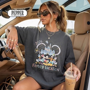 May include: Dark grey t-shirt with a Disney-themed graphic. The design includes Mickey Mouse, Minnie Mouse, Donald Duck, and other characters, a castle, and the text "2026 FAMILY VACATION". The word "PEPPER" is in a white rectangle in the top left corner.