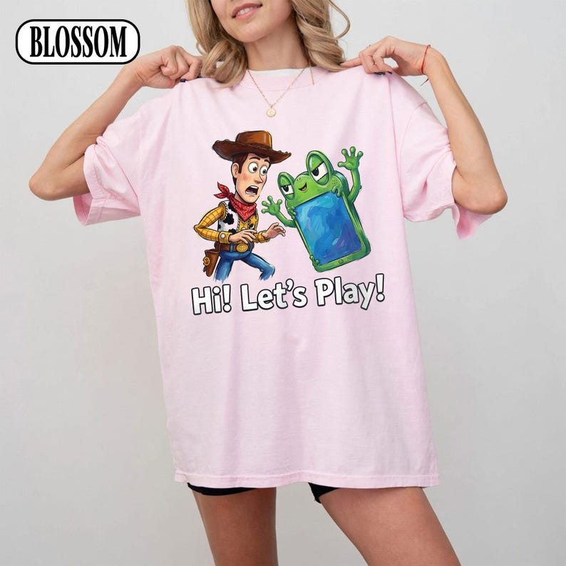May include: A light pink t-shirt featuring a graphic print of a cowboy character in a brown hat and a green frog character holding a blue tablet. The text "Hi! Let's Play!" is printed in black below the characters. The word "BLOSSOM" is in the corner.