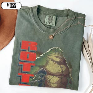 Puede incluir: Camiseta verde Comfort Colors con un gr&aacute;fico de una criatura grande y verde y la palabra "ROTT" en rojo. La camiseta tiene cuello redondo y una peque&ntilde;a etiqueta que dice "Comfort Colors". Tambi&eacute;n se ve un collar dorado con un colgante.