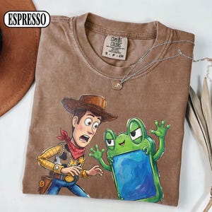 May include: An espresso-coloured short-sleeve t-shirt featuring a graphic print of a cowboy character with a brown hat and red bandana standing next to a green frog character holding a blue rectangular object. The shirt has a crew neckline.