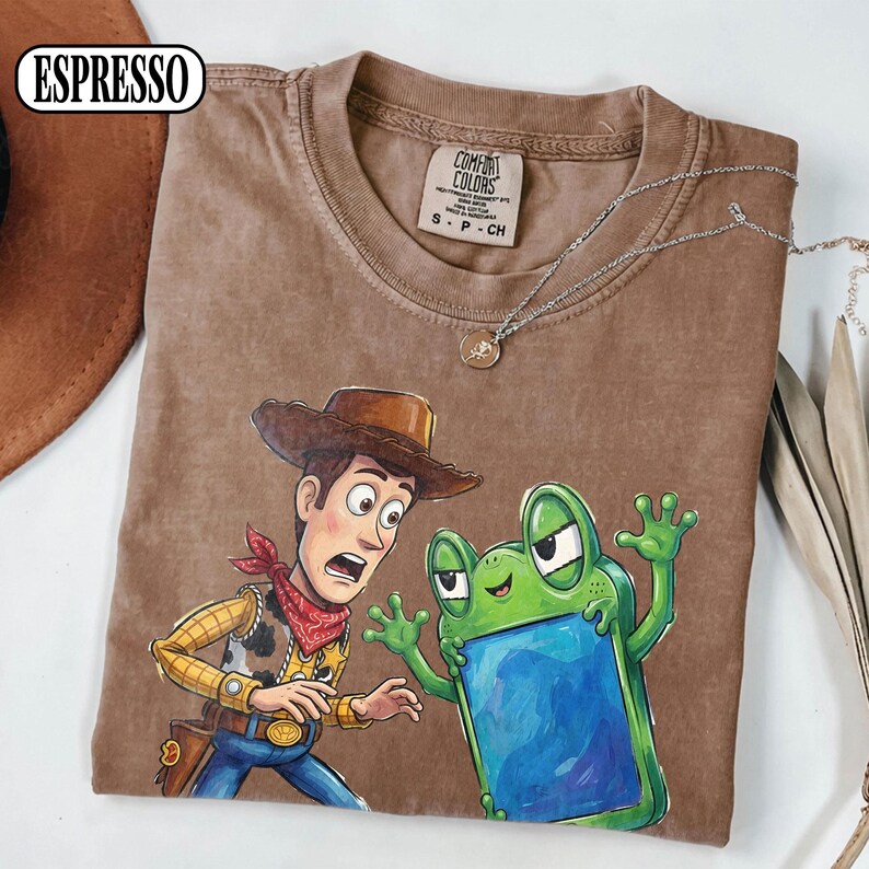May include: An espresso-coloured short-sleeve t-shirt featuring a graphic print of a cowboy character with a brown hat and red bandana standing next to a green frog character holding a blue rectangular object. The shirt has a crew neckline.