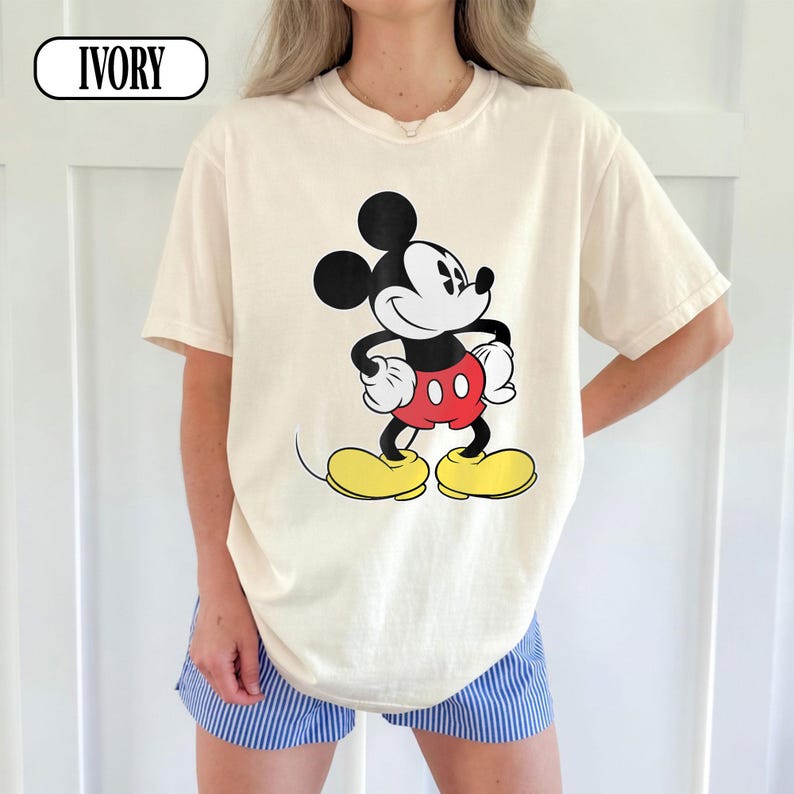 May include: An ivory short-sleeve t-shirt featuring a large graphic print of Mickey Mouse on the front. The character is depicted in black, white, red, and yellow. The text IVORY is printed in a black rectangular box in the upper left corner.