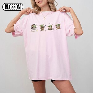 May include: A light pink t-shirt featuring a graphic of a character from a popular science fiction series. The graphic shows the character in four different poses. The word "cool" is printed on the shirt. The brand name "BLOSSOM" is printed on the top left.