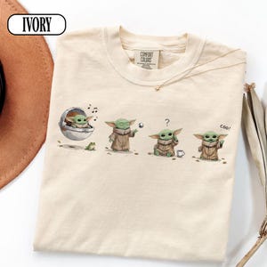 May include: Ivory-colored t-shirt featuring a series of illustrations of a green alien character in a brown cloak. The character is depicted in various poses, including one in a floating pod, and another with a question mark above its head. The word "coo!" is also present.