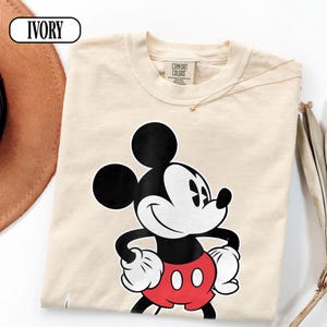 May include: An ivory-colored short-sleeve t-shirt featuring a graphic print of Mickey Mouse. The character is depicted in black, white, and red, with hands on hips. The shirt has a Comfort Colors brand label at the inner neckline and the word IVORY.