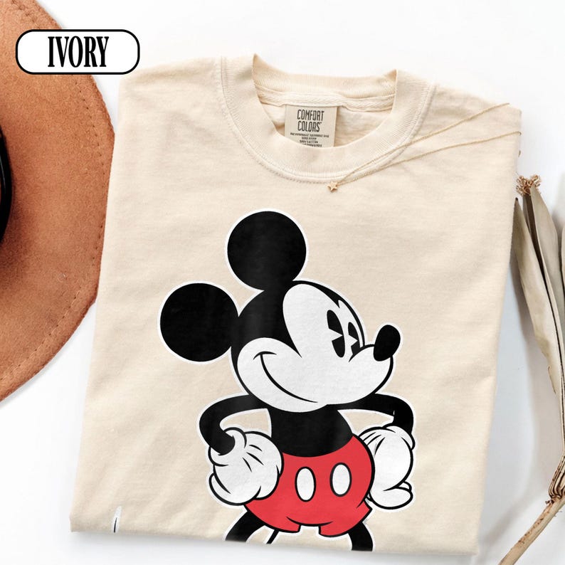 May include: An ivory-colored short-sleeve t-shirt featuring a graphic print of Mickey Mouse. The character is depicted in black, white, and red, with hands on hips. The shirt has a Comfort Colors brand label at the inner neckline and the word IVORY.