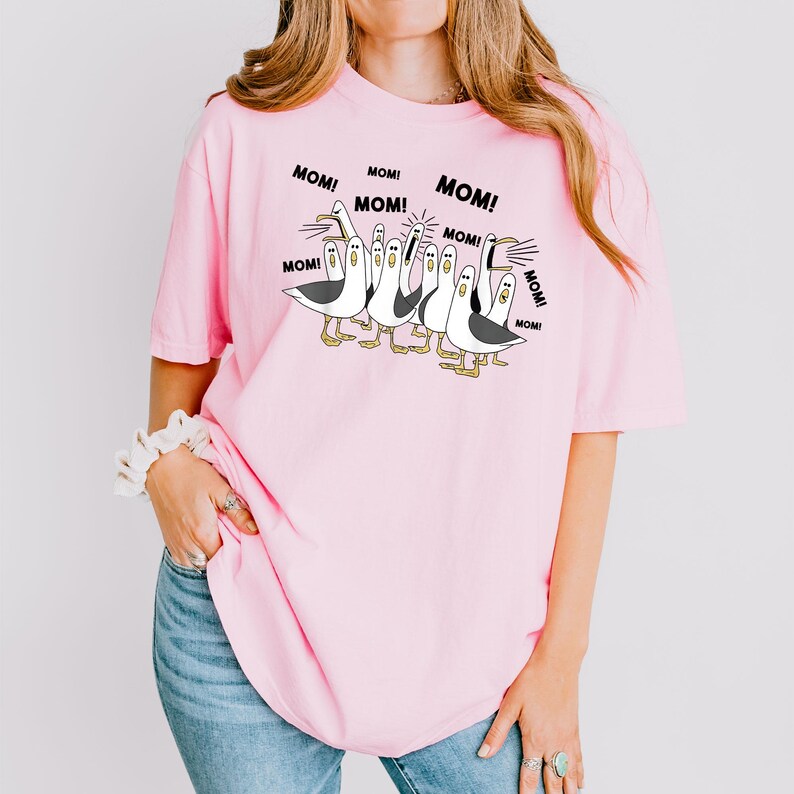 以下が含まれることがあります： ピンク色のTシャツ。カモメの漫画イラストが特徴で、各カモメの上に「MOM！」の文字が描かれています。カモメは白黒で、足とくちばしは黄色です。シャツは明るいピンク色です。