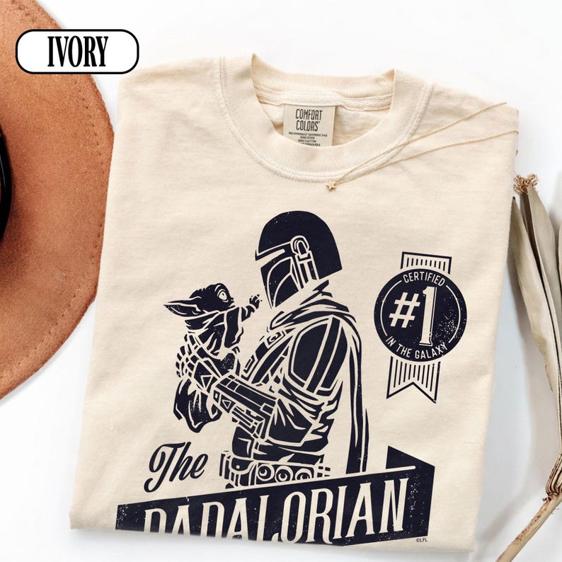 Puede incluir: Camiseta color marfil con un gr&aacute;fico negro de The Mandalorian sosteniendo a Grogu. El texto dice "The Dadalorian" y "Certified #1 in the Galaxy." Marca Comfort Colors.