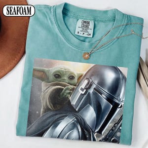 Puede incluir: Camiseta verde espuma de mar con un gr&aacute;fico de The Mandalorian y Grogu. La camiseta tiene cuello redondo y mangas cortas. Una etiqueta Comfort Colors es visible. Un collar dorado con un colgante est&aacute; drapeado sobre la camiseta.