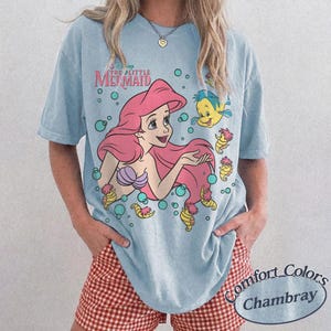 May include: A light blue short-sleeve t-shirt featuring a graphic of Ariel from The Little Mermaid with pink hair, a purple seashell top, and the yellow fish Flounder. The design includes small bubbles and sea creatures in a cartoon style.