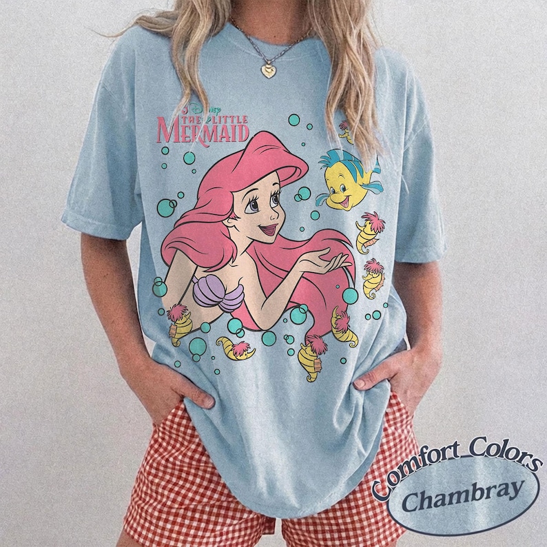 May include: A light blue short-sleeve t-shirt featuring a graphic of Ariel from The Little Mermaid with pink hair, a purple seashell top, and the yellow fish Flounder. The design includes small bubbles and sea creatures in a cartoon style.