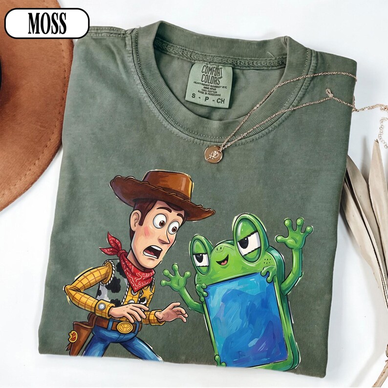 May include: A moss green t-shirt featuring a graphic print of a cowboy character with a brown hat and red bandana standing next to a green frog character holding a blue rectangular object. The shirt has a crew neckline and a Comfort Colors brand label.