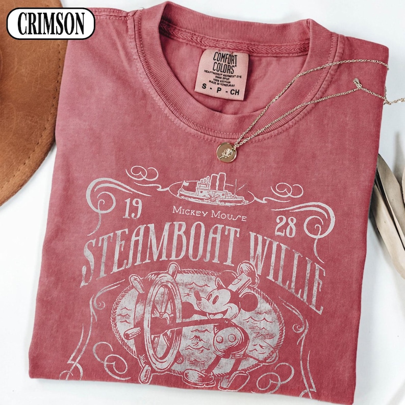 May include: A crimson red short-sleeve t-shirt featuring a white graphic print of Steamboat Willie with the text 1928 Mickey Mouse Steamboat Willie. The shirt has a crew neckline and a Comfort Colors brand label printed on the inside of the collar.