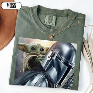 Puede incluir: Camiseta verde oliva con un gr&aacute;fico de The Mandalorian y Grogu. La camiseta tiene cuello redondo y mangas cortas. Un collar dorado con un colgante est&aacute; drapeado sobre la camiseta. La palabra "MOSS" est&aacute; en un &oacute;valo blanco en la esquina superior izquierda.