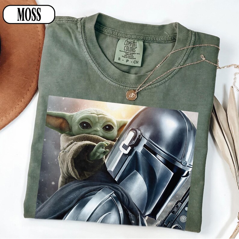 Puede incluir: Camiseta verde oliva con un gr&aacute;fico de The Mandalorian y Grogu. La camiseta tiene cuello redondo y mangas cortas. Un collar dorado con un colgante est&aacute; drapeado sobre la camiseta. La palabra "MOSS" est&aacute; en un &oacute;valo blanco en la esquina superior izquierda.