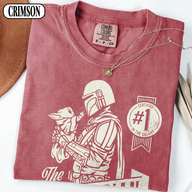 Puede incluir: Camiseta roja carmes&iacute; con un gr&aacute;fico de un personaje mandaloriano sosteniendo una peque&ntilde;a criatura. La camiseta incluye el texto "Certified #1 in the Galaxy" y "The". Un collar dorado con un colgante est&aacute; sobre la camiseta.
