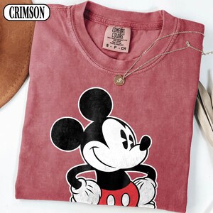 May include: A crimson t-shirt featuring a graphic of Mickey Mouse with black ears, a white face, and red shorts. The shirt has a Comfort Colors label at the neck with text reading S P CH. A thin gold-toned chain necklace rests on the fabric.