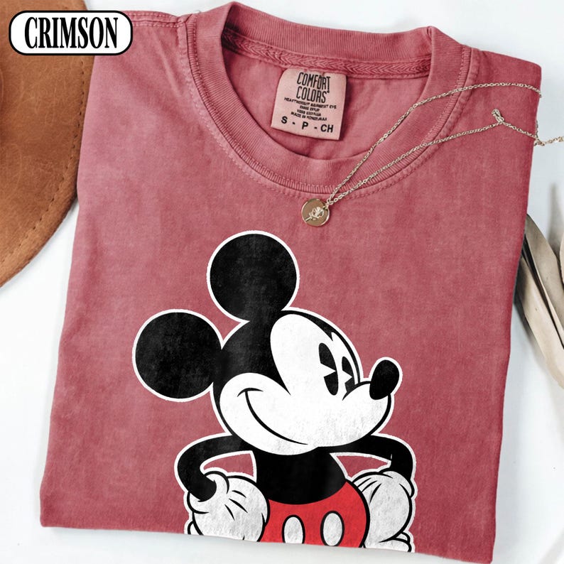 May include: A crimson t-shirt featuring a graphic of Mickey Mouse with black ears, a white face, and red shorts. The shirt has a Comfort Colors label at the neck with text reading S P CH. A thin gold-toned chain necklace rests on the fabric.